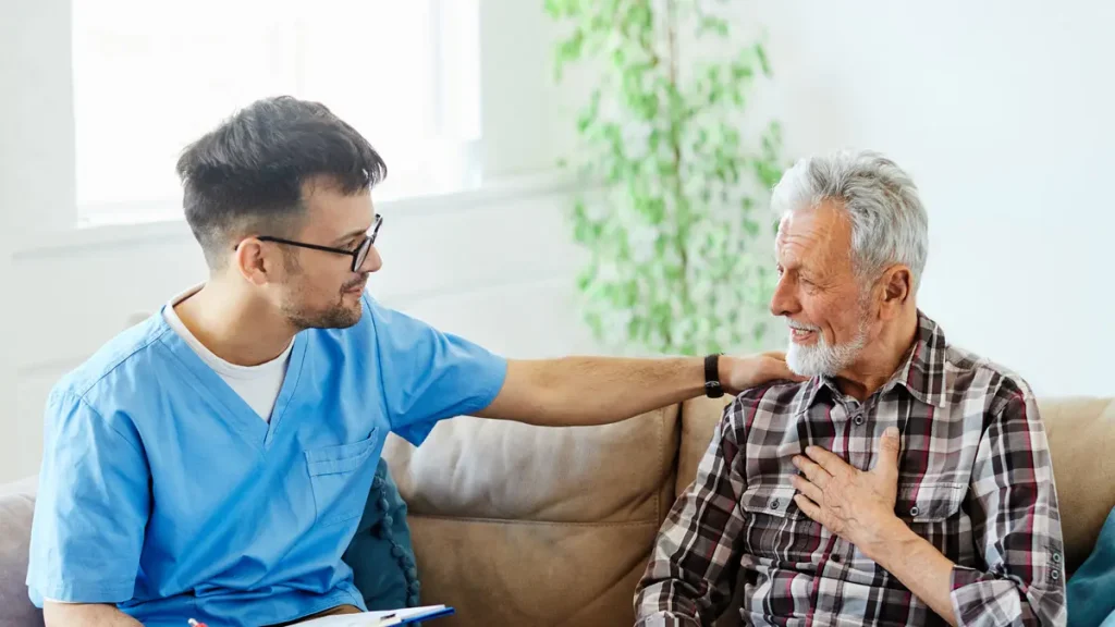 A Kindcare at Bristol caregiver in blue scrubs comforts an older man on a couch in a bright living room.